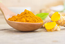 site-industries-cosmetiques Une cuillère en bois remplie de poudre de curcuma moulu se trouve au premier plan, avec des tranches de racine de curcuma fraîche et un fond vert feuillu sur une surface en bois clair.
