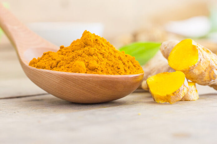 site-industries-cosmetiques Une cuillère en bois remplie de poudre de curcuma moulu se trouve au premier plan, avec des tranches de racine de curcuma fraîche et un fond vert feuillu sur une surface en bois clair.