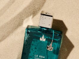 site-industries-cosmetiques 一个标有 "LE REM LIMITED EDITION 1996 REMINISCENCE JUAN-LES-PINS "字样的蓝色长方形香水瓶放在沙地上，瓶上有水滴，瓶盖上用蓝色绳索系着一个小银坠。.
