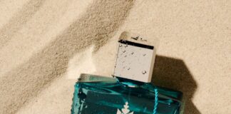 site-industries-cosmetiques Un flacon de parfum rectangulaire bleu étiqueté "LE REM LIMITED EDITION 1996 REMINISCENCE JUAN-LES-PINS" repose sur du sable, avec des gouttes d'eau sur le flacon et une petite breloque en argent attachée au bouchon par un cordon bleu.