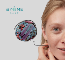 site-industries-cosmetiques Grande plano de uma mulher a tocar na sua bochecha, com uma vista ampliada que mostra bactérias coloridas na sua pele. O logótipo da Byome Labs aparece no canto superior esquerdo.