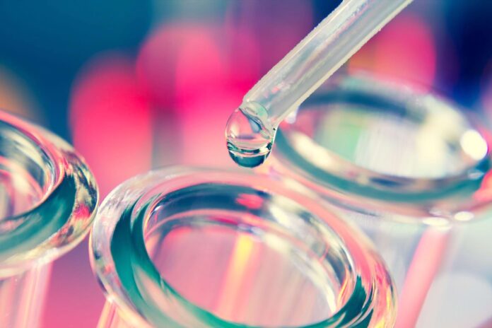 site-industries-cosmetiques Primer plano de una pipeta dispensando una gota de líquido en un tubo de ensayo de vidrio, con otros dos tubos de ensayo visibles. El fondo está difuminado, con tonos rosas y azules.
