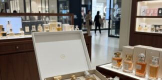 site-industries-cosmetiques 在一家商店的木桌上展示一个打开的盒子中的豪华香水瓶，周围散落着白色花瓣和玻璃珠。背景中可以看到装有其他香水瓶的货架。.