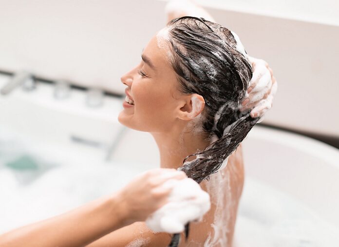 site-industries-cosmetiques Uma mulher lava o cabelo numa banheira, sorrindo e fechando os olhos. O seu cabelo está coberto de espuma de champô e as suas mãos massajam o couro cabeludo. O fundo é brilhante e desfocado.