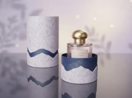 site-industries-cosmetiques 一个带金色瓶塞的玻璃香水瓶被装在一个打开的圆柱形盒子中，盒子上装饰有花卉图案和波浪形的蓝色装饰。与之相匹配的盒盖立在盒子旁边的反光表面上。.