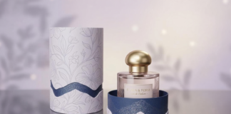 site-industries-cosmetiques 一个带金色瓶塞的玻璃香水瓶被装在一个打开的圆柱形盒子中，盒子上装饰有花卉图案和波浪形的蓝色装饰。与之相匹配的盒盖立在盒子旁边的反光表面上。.