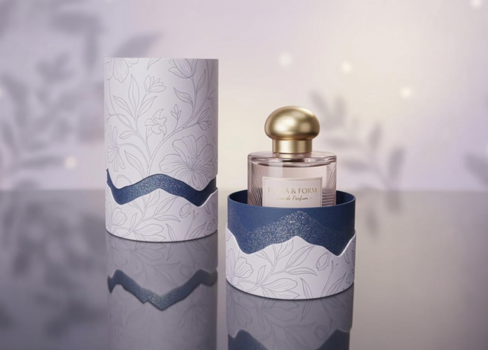 site-industries-cosmetiques 一个带金色瓶塞的玻璃香水瓶被装在一个打开的圆柱形盒子中，盒子上装饰有花卉图案和波浪形的蓝色装饰。与之相匹配的盒盖立在盒子旁边的反光表面上。.