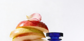 site-industries-cosmetiques 在白色背景下，一个蓝色玻璃瓶旁边竖立着一堆切片的苹果、梨、香蕉、草莓和无花果，上面点缀着薄荷叶和玫瑰花瓣。.