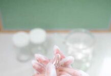 site-industries-cosmetiques Une paire de mains aux ongles vernis de couleur claire est lavée avec du savon, créant une mousse épaisse et des bulles. L'arrière-plan montre un verre d'eau et des récipients blancs flous sur un comptoir.