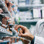 site-industries-cosmetiques 两个穿着白大褂的人站在摆满药品的货架前。其中一人拿着药瓶，另一人在一旁看着。背景似乎是药房或医疗用品室。.