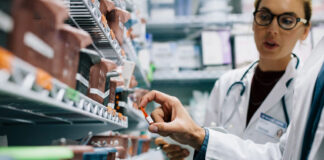 site-industries-cosmetiques Deux personnes en blouse se tiennent devant des étagères remplies de médicaments. L'une d'entre elles tient un flacon de pilules tandis que l'autre regarde. Le décor semble être celui d'une pharmacie ou d'une salle de fournitures médicales.