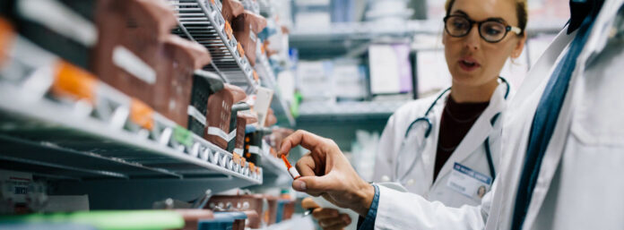 site-industries-cosmetiques Two people in lab coats stand in front of shelves filled with medicines. One holds a bottle of pills while the other looks on. The setting appears to be that of a pharmacy or medical supply room.