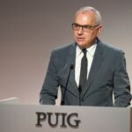 site-industries-cosmetiques 一名戴眼镜、穿灰色西装、打黑色领带的男子站在刻有 "PUIG "字样的讲台后面，在浅色背景下对着麦克风讲话。.