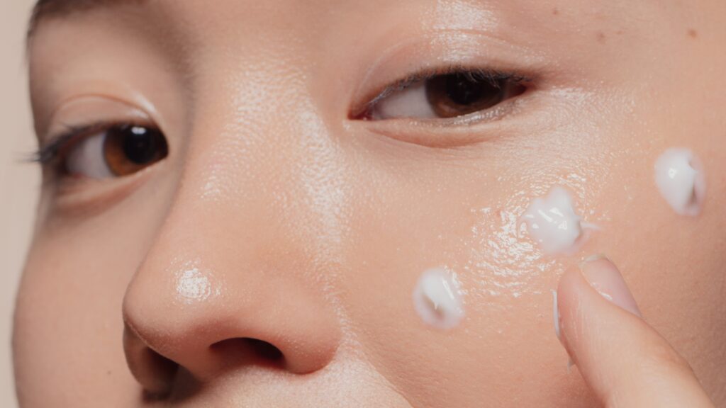 site-industries-cosmetiques Close-up of a person applying small dots of white cream to her cheek with her finger. The smooth, well-lit complexion highlights the benefits of structured skin, while the person's eye is partially visible.
