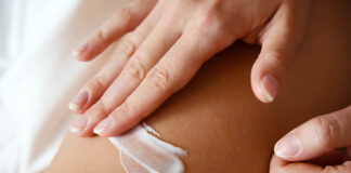 site-industries-cosmetiques Gros plan sur la main d'une personne appliquant un filet de lotion ou de crème blanche sur sa peau nue, probablement sur la jambe ou le bras. L'accent est mis sur la main et sur la crème appliquée.