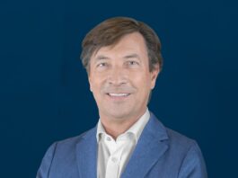site-industries-cosmetiques 一名身穿蓝色西装外套和白色衬衫的黑发男子站在深蓝色背景前，面带微笑，双臂松松地交叉着。.