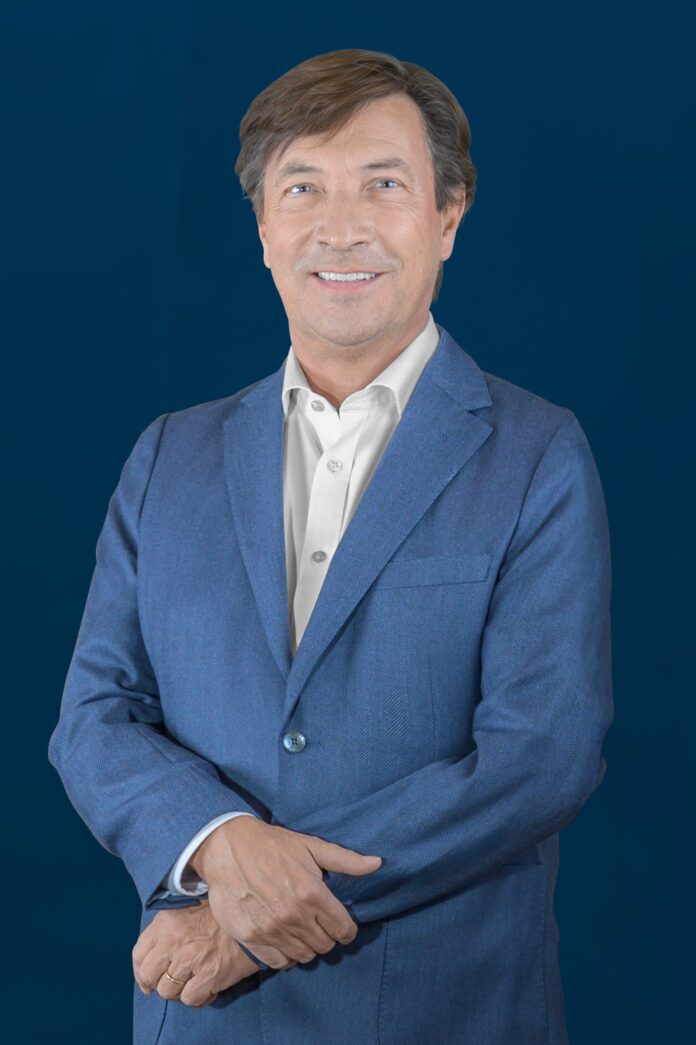 site-industries-cosmetiques Un homme aux cheveux bruns portant une veste de costume bleue et une chemise blanche se tient devant un fond bleu foncé uni, souriant et les bras vaguement croisés.