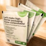 Basf élargit l’offre ecovio pour les emballages flexibles site-industries-cosmetiques Présentation d'échantillons d'emballages compostables de la marque BASF dans un conteneur transparent sur une surface en bois, avec un texte faisant la promotion d'emballages compostables à domicile à haute barrière et présentant des avantages écologiques.