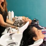 欧莱雅基金会推出第二家流动护理沙龙 site-industries-cosmetiques 两个人坐在发廊的洗头台前,头向后仰,发型师正在为其中一人洗头。两位顾客看起来都很放松。背景中可以看到美发产品的瓶子。.