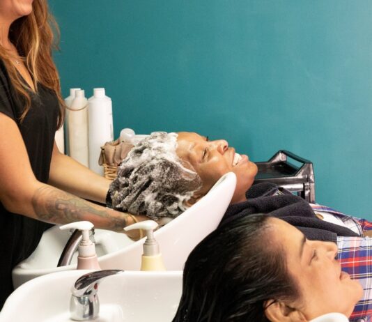 site-industries-cosmetiques Two people sit at the salon's wash stations, heads tilted back, while a stylist washes the hair of one of them. Both customers look relaxed. Bottles of hair care products are visible in the background.