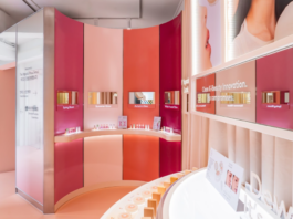site-industries-cosmetiques Un espace de présentation moderne dédié aux produits de beauté se compose de murs incurvés dans des tons de rose et d'orange, d'étagères proposant des produits de soin, d'écrans numériques au-dessus de chaque rayon et d'affiches promotionnelles accrochées au mur. Un éclairage vif et homogène met l'ensemble en valeur.