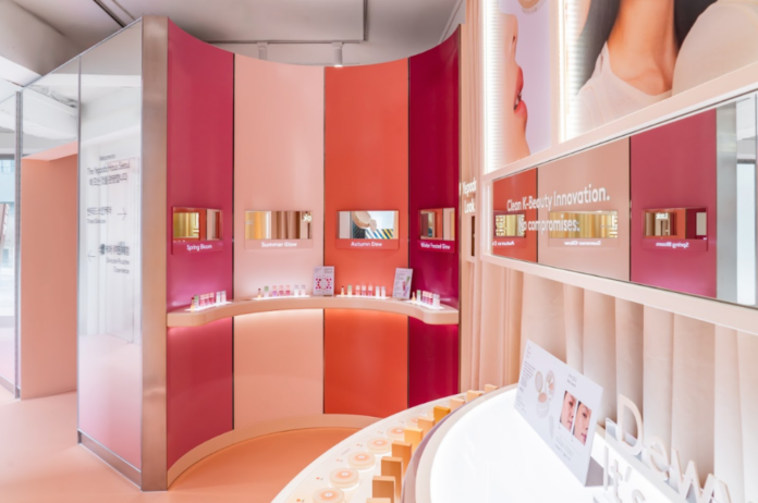 site-industries-cosmetiques Un espace de présentation moderne dédié aux produits de beauté se compose de murs incurvés dans des tons de rose et d'orange, d'étagères proposant des produits de soin, d'écrans numériques au-dessus de chaque rayon et d'affiches promotionnelles accrochées au mur. Un éclairage vif et homogène met l'ensemble en valeur.