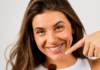 site-industries-cosmetiques Una mujer de pelo largo y castaño, con un top blanco sin mangas, sonríe y sostiene una brocha de maquillaje, de cara a la cámara, sobre un fondo claro y liso.
