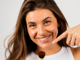 site-industries-cosmetiques Una mujer de pelo largo y castaño, con un top blanco sin mangas, sonríe y sostiene una brocha de maquillaje, de cara a la cámara, sobre un fondo claro y liso.