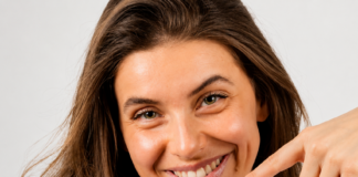 site-industries-cosmetiques Una mujer de pelo largo y castaño, con un top blanco sin mangas, sonríe y sostiene una brocha de maquillaje, de cara a la cámara, sobre un fondo claro y liso.