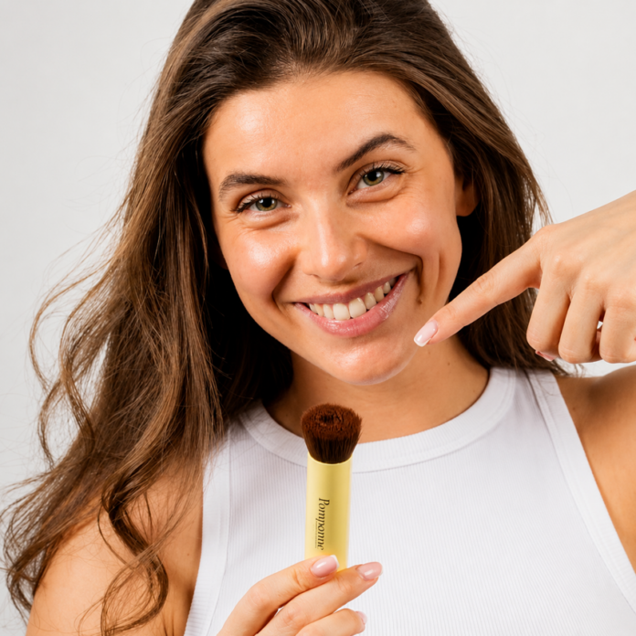 site-industries-cosmetiques A woman with long brown hair, wearing a sleeveless white top, smiles and shows a make-up brush she is holding, facing the camera, against a light, plain background.