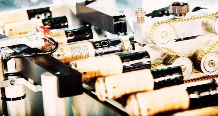 site-industries-cosmetiques Vue rapprochée d'une machine appliquant des étiquettes sur des bouteilles de vin munies de bouchons en liège sur une ligne de production automatisée. Les bouteilles sont déplacées par des rouleaux et des engrenages dans le cadre du processus d'étiquetage.