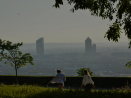 site-industries-cosmetiques Deux personnes marchent le long d'un sentier bordé d'herbe et d'arbres, avec en arrière-plan la silhouette floue de la ville et deux gratte-ciel qui se détachent clairement. La scène semble se dérouler tôt le matin ou en fin d'après-midi.