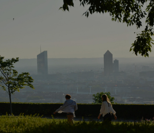 site-industries-cosmetiques Deux personnes marchent le long d'un sentier bordé d'herbe et d'arbres, avec en arrière-plan la silhouette floue de la ville et deux gratte-ciel qui se détachent clairement. La scène semble se dérouler tôt le matin ou en fin d'après-midi.