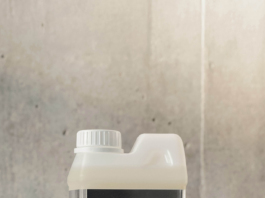 site-industries-cosmetiques Un récipient en plastique muni d'un bouchon à vis blanc repose sur une surface en béton. L'étiquette porte l'inscription « EAU DE LESSIVE ÉDITION CONCRÈTE bdlk » en lettres blanches sur fond noir. On aperçoit le mur en béton à l'arrière-plan.