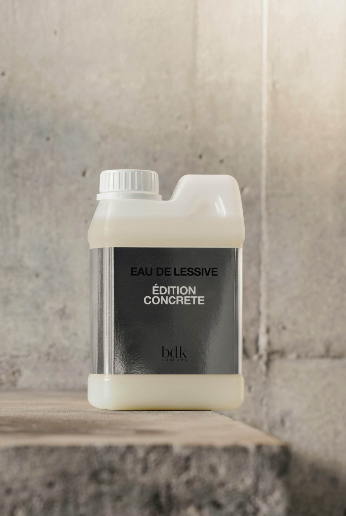 site-industries-cosmetiques A plastic container with a white screw-top cap sits on a concrete surface. The label reads «EAU DE LESSIVE ÉDITION CONCRÈTE bdlk» in white letters on a black background. The concrete wall is visible in the background.