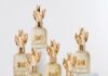 site-industries-cosmetiques Seis frascos de perfume em vidro, com tampas douradas em forma de coroa, estão dispostos sobre pedestais brancos. Cada frasco apresenta uma textura estriada e um rótulo dourado no qual estão impressos algarismos romanos e nomes. O fundo é de um branco liso.