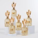 El Grupo TNT fabrica en una sola pieza la tapa de zamak de la colección «Essences Précieuses».» site-industries-cosmetiques Seis frascos de perfume de cristal, provistos de tapones dorados con forma de corona, están dispuestos sobre peanas blancas. Cada frasco presenta una textura acanalada y una etiqueta dorada en la que están impresos números romanos y nombres. El fondo es de un blanco liso.
