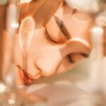 site-industries-cosmetiques Primer plano de una persona con los ojos cerrados, con el rostro apoyado sobre una superficie, mientras una luz suave y las sombras proyectadas por objetos de cristal dibujan motivos sobre su piel.