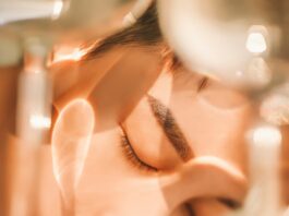 site-industries-cosmetiques Gros plan sur une personne aux yeux fermés, le visage posé sur une surface, tandis qu’une lumière douce et les ombres projetées par des objets en verre dessinent des motifs sur sa peau.