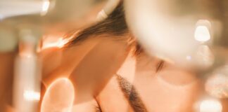 site-industries-cosmetiques Grande plano de uma pessoa com os olhos fechados, com o rosto apoiado numa superfície, enquanto uma luz suave e as sombras projetadas por objetos de vidro desenham padrões na sua pele.