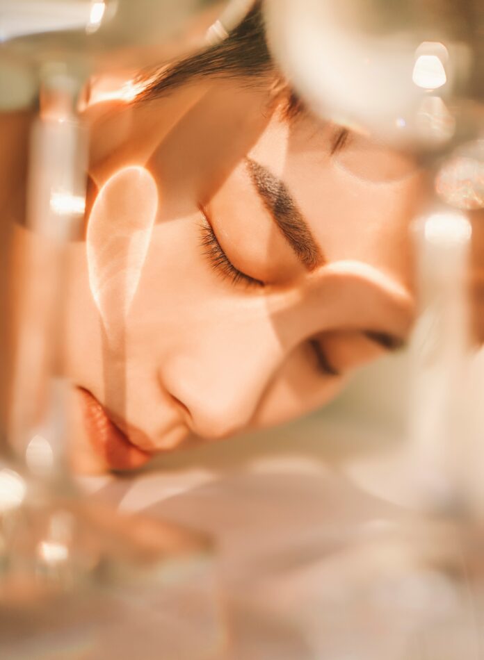 site-industries-cosmetiques Grande plano de uma pessoa com os olhos fechados, com o rosto apoiado numa superfície, enquanto uma luz suave e as sombras projetadas por objetos de vidro desenham padrões na sua pele.