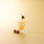 site-industries-cosmetiques 一瓶装有SO'BIO étic眼部精华液的透明玻璃瓶，配有金色和白色的滴管，置于浅棕色台面上，旁边放着两颗棕色的阿甘坚果，整体背景为相配的米色。.