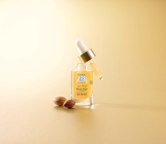 site-industries-cosmetiques Um frasco de vidro transparente contendo o sérum para os olhos SO'BIO étic, equipado com um conta-gotas dourado e branco, pousado sobre uma superfície castanha clara, com duas nozes de argão castanhas ao lado, tudo isto sobre um fundo bege a condizer.
