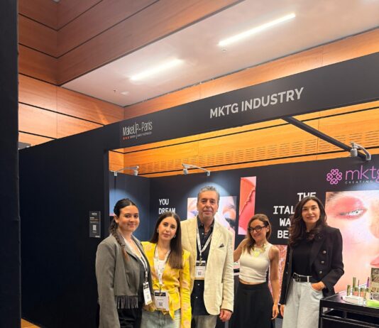 site-industries-cosmetiques Quatre personnes se tiennent debout et posent pour une photo devant un stand noir portant l'inscription « MKTG INDUSTRY », dans ce qui semble être un salon professionnel ou une exposition. Le stand présente des marques de maquillage et du matériel promotionnel.