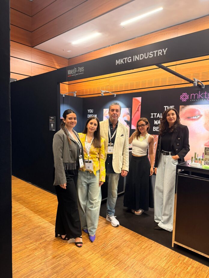site-industries-cosmetiques Four people are standing and posing for a photo in front of a black booth bearing the inscription «MKTG INDUSTRY,» at what appears to be a trade show or exhibition. The booth features makeup brands and promotional materials.