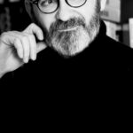 Frank Sorbier lanza Sorbier Parfums, una colección de alta perfumería dividida en tres «capítulos» site-industries-cosmetiques Retrato en blanco y negro de una persona con barba y bigote, que lleva gafas y un jersey de cuello alto oscuro, con la mano apoyada en la cara y la mirada pensativa dirigida hacia el horizonte. Se ven cajas de cartón al fondo.