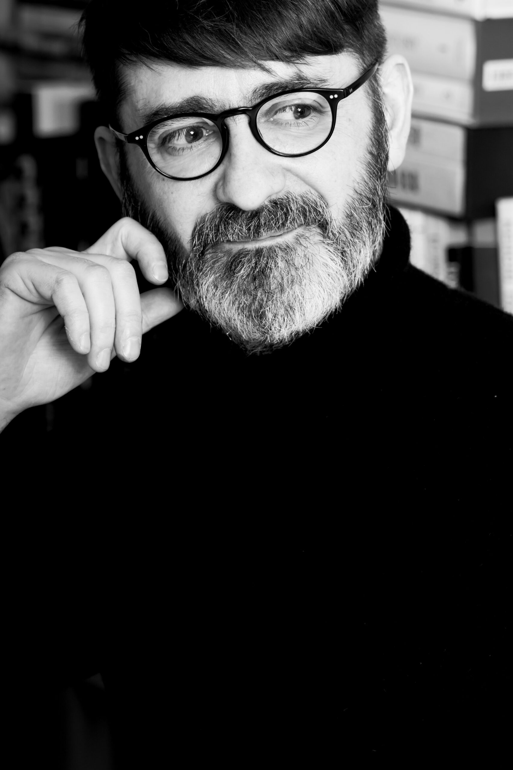 site-industries-cosmetiques Portrait en noir et blanc d'une personne barbu et moustachu, portant des lunettes et un col roulé foncé, la main posée sur le visage, le regard pensif tourné vers l'horizon. On aperçoit des cartons à l'arrière-plan.