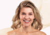 Liftilience for textured hair: hair efficacy data site-cosmetics-industries A woman with shoulder-length blonde hair smiles at the camera against a light-colored background adorned with beige wavy lines. She has fair skin and is photographed from the head down to her shoulders.