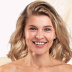 Liftilience para cabelos texturados: dados sobre a eficácia capilar site-industries-cosmetiques Uma mulher de cabelo loiro até aos ombros sorri para a câmara diante de um fundo claro decorado com linhas onduladas em tons de bege. Tem a pele clara e é fotografada da cabeça aos ombros.