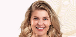 Liftilience para cabellos con textura: datos sobre la eficacia capilar site-industries-cosmetiques Una mujer de cabello rubio medio largo sonríe a la cámara ante un fondo claro decorado con líneas onduladas de color beige. Tiene la tez clara y aparece fotografiada de la cabeza a los hombros.